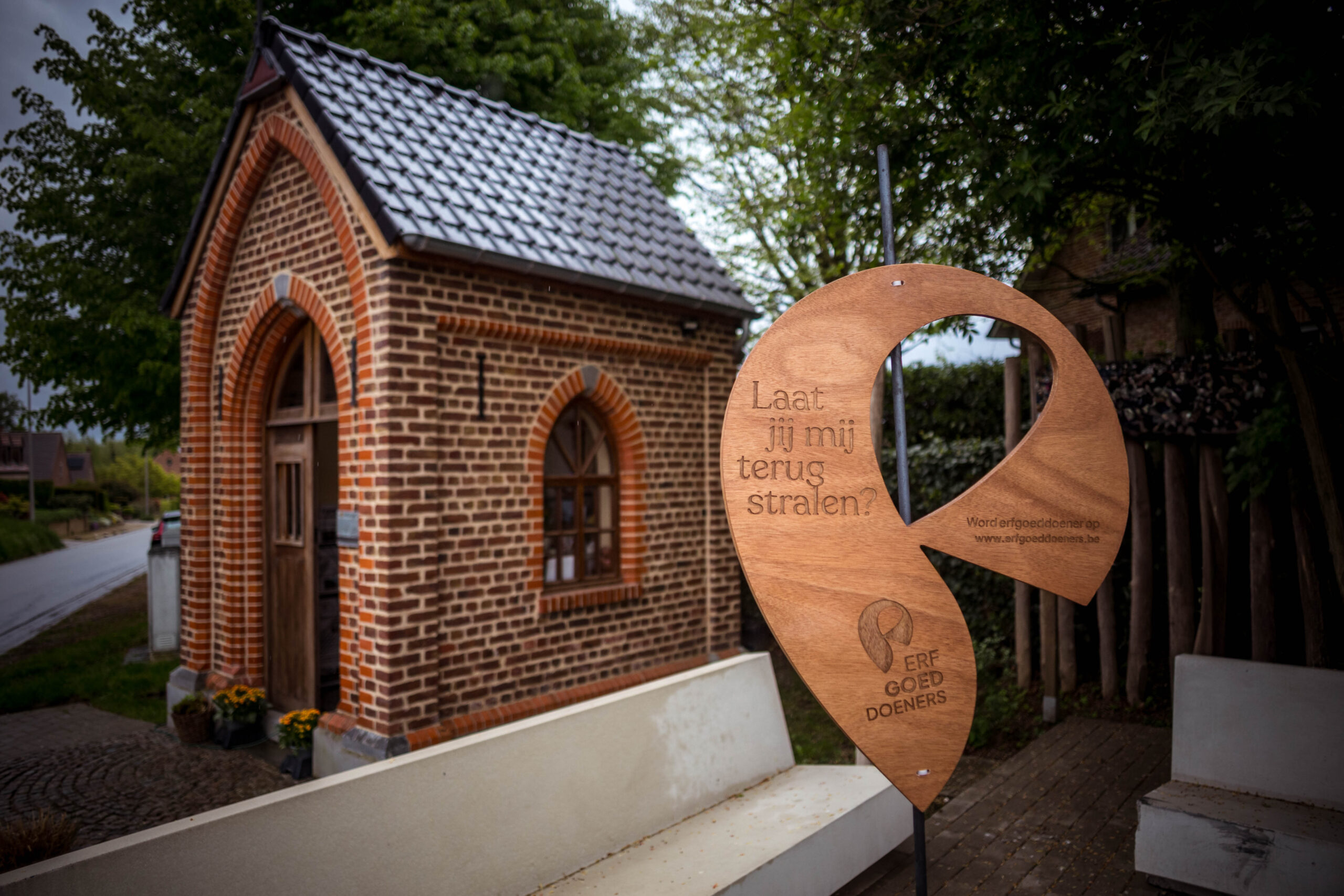 Bakstenen kapel met erfgoedbord, bloemen ernaast.